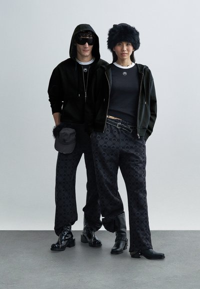 Melnā kapuci ar rāvējslēdzēju, kombinēta ar piegulošu melnu topu un rakstainām tumšām biksēm. Aksesuāri: melna cepure un kažokādas cepure. Melni zābaki.