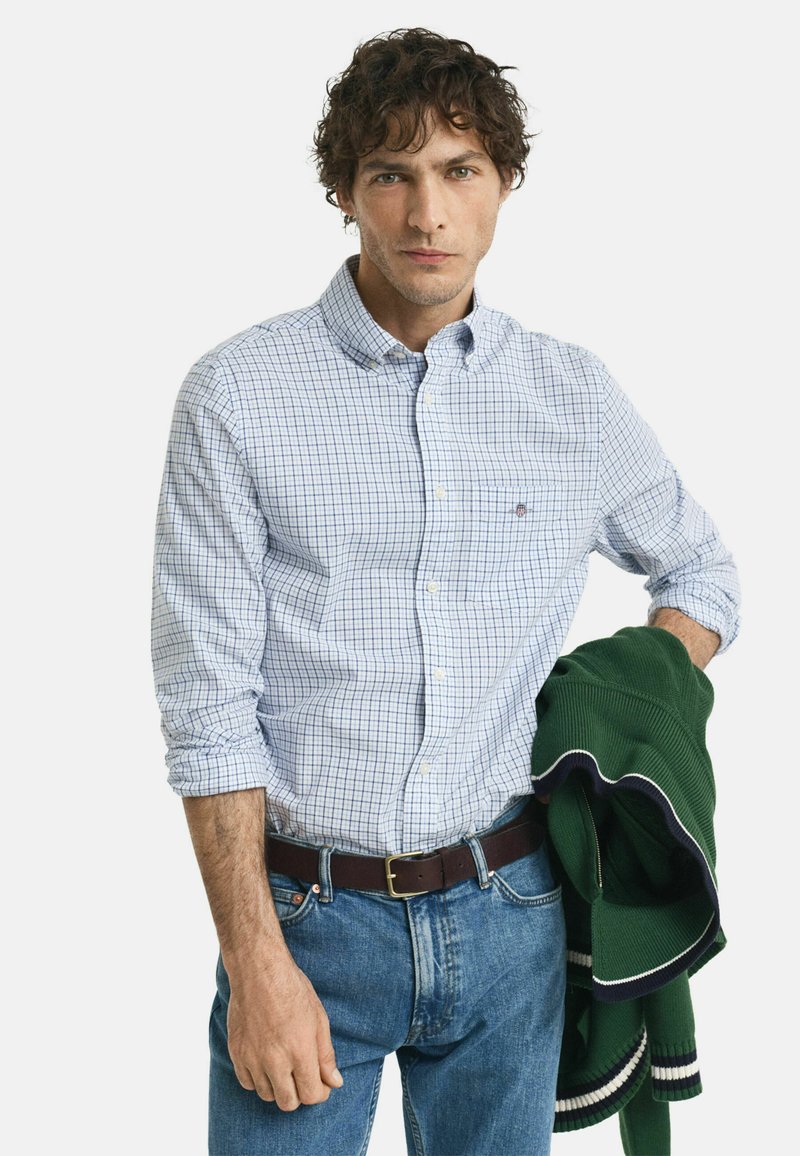 Mann mit lockigem Haar, der ein blau kariertes Hemd und Jeans trägt, hält einen grünen Pullover mit weißem und marineblauem Besatz über einem Arm.