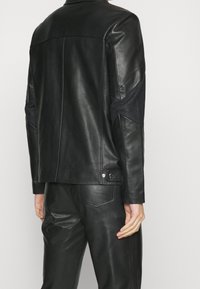 Veste en cuir noir avec des accents de tissu sombre ton sur ton sur les manches, coupe ajustée, et détails à pression au col et à l'ourlet. Texture lisse.