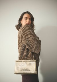 Goldfarbener metallischer Handtasche mit glatter Textur und rechteckiger Form, ausgestattet mit einem kurzen Riemen. Das Modell trägt einen braunen, flauschigen Pullover und dunkle Hosen.