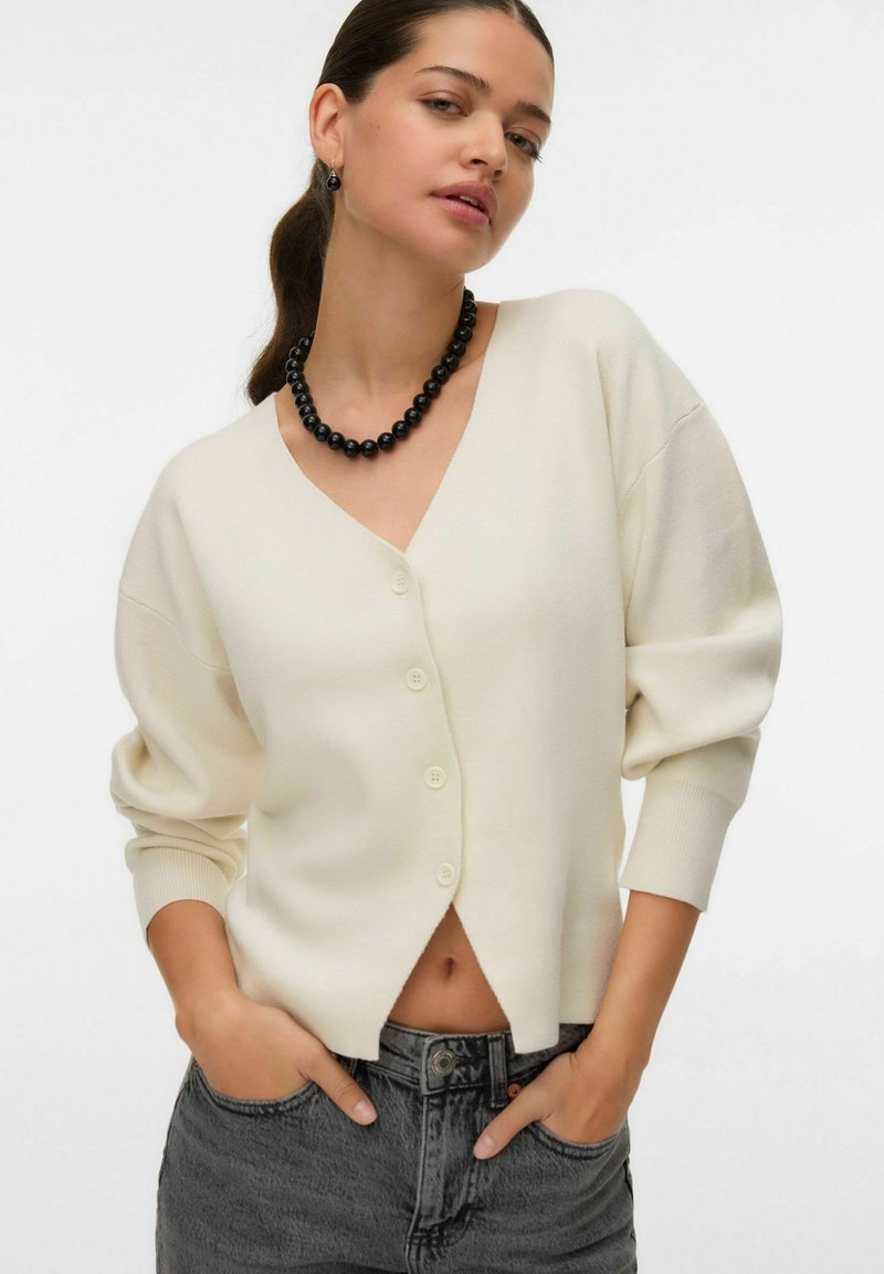 Romige gebreide cardigan met een V-hals, knoopsluiting en cropped lengte. Gecombineerd met high-waisted donkere jeans en een zwarte kralenketting.