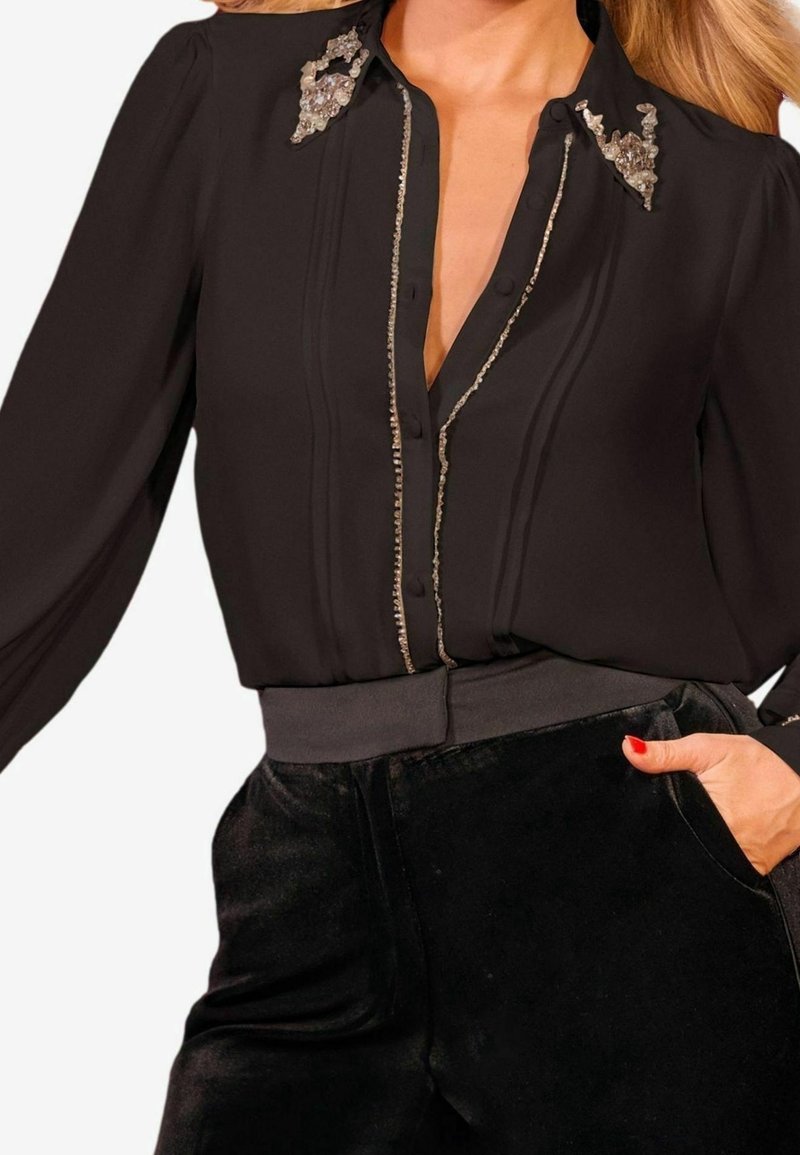 Svart blus med djup V-ringning, prydd med silverdetaljer på kragen och en knapprad framtill; kombinerad med svarta texturerade byxor.