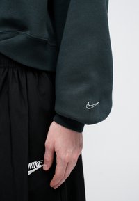 Tumši zaļš džemperis ar ribotām piedurknēm un mazu balto izšūto Nike logo uz piedurknes, kombinēts ar melnu apģērbu.