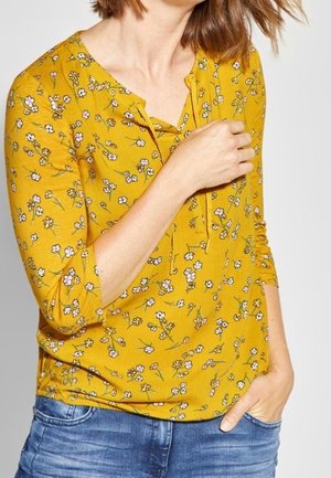 Femme portant un haut à manches longues à fleurs jaune moutarde avec un col à nouer et un jean bleu, une main dans la poche et l'autre près de sa poitrine.