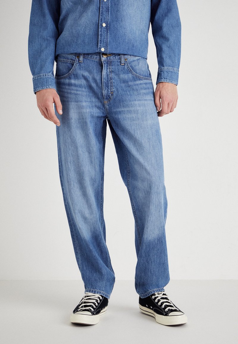 Jeans in denim azzurro chiaro con una vestibilità comoda, chiusura con bottone, cinque tasche e cuciture in contrasto, abbinati a sneakers nere.