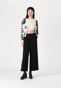 Black and white patterned cardigan over a cropped white top, paired with high-waisted black wide-leg trousers and chunky black shoes.