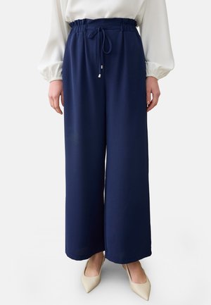 Frau trägt marineblaue, weit geschnittene Hose mit Kordelzug in der Taille, weiße Langarmbluse und beige spitze Schuhe, steht vor weißem Hintergrund.