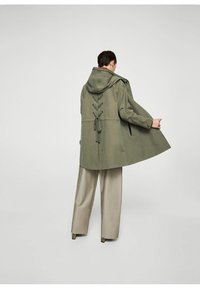 Veste anorak vert olive avec une capuche, lacets ajustables à l'arrière et poches latérales, associée à un pantalon large beige clair.