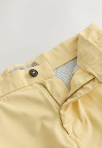 Pantalon jaune clair avec fermeture éclair partiellement ouverte, bouton noir et doublure intérieure visible à rayures ceinturant la taille.
