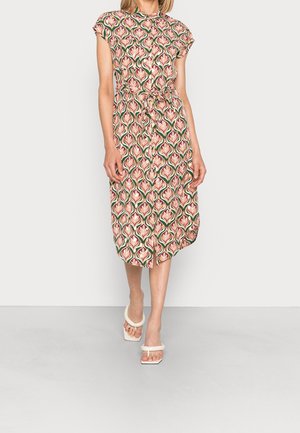 Kvinde iført midlang kjole med pink og grøn blomstermønster, korte ærmer, taljebånd og hvide sandaler med hæl, der går fremad.