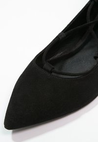 Zapato plano de ante negro con punta afilada y correas entrelazadas. Forro interior suave, diseño minimalista y sutilezas en los detalles de costura.