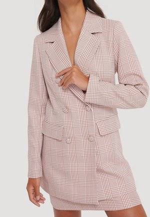 Frau trägt ein pink-weiß kariertes, zweireihiges Blazer-Kleid mit Revers und Klappentaschen und richtet mit einer Hand den Kragen.