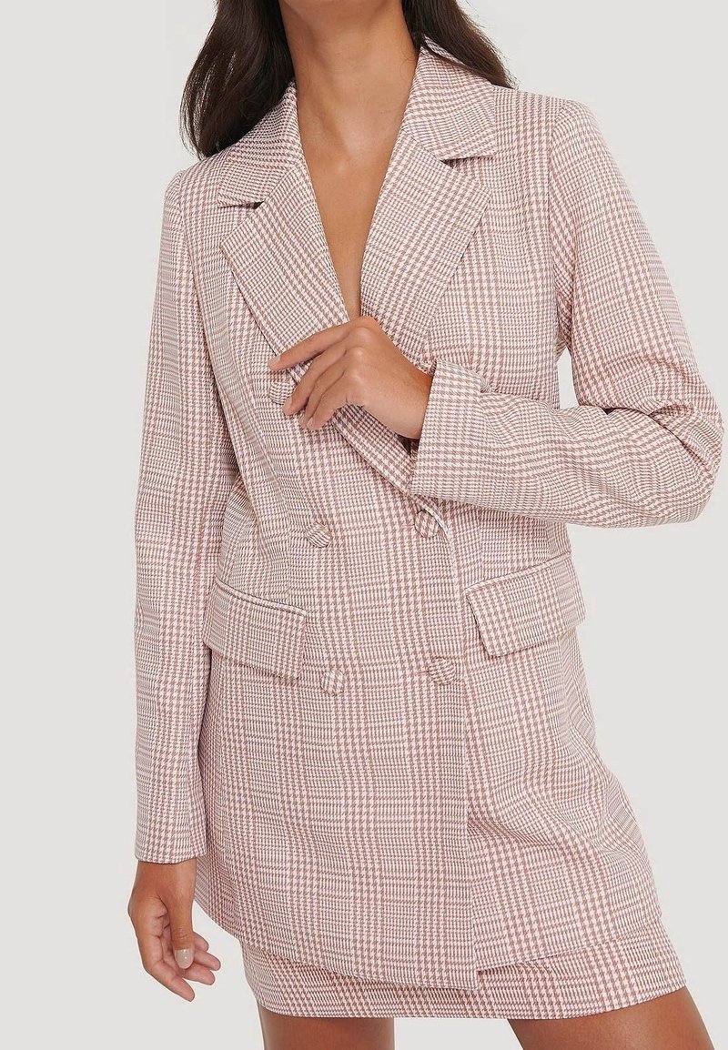 Femme portant une robe blazer à carreaux rose et blanc, croisée, avec revers et poches à rabat, ajustant le col d'une main.