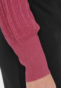 Haut à manches longues avec une manche rose texturée et côtelée présentant des motifs verticaux et un fil métallique. Le tissu noir contraste avec la manche.