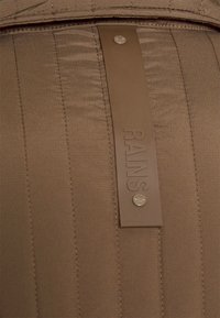 Brunt quiltat tyg med en rektangulär läderetikett präglad med "RAINS" och en silverknappdetalj.