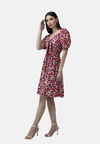 Vestido rojo con flores, mangas abullonadas, cintura ceñida, dobladillo a la altura de la rodilla y sandalias blancas con tacón delgado. Tejido ligero con patrones vibrantes.