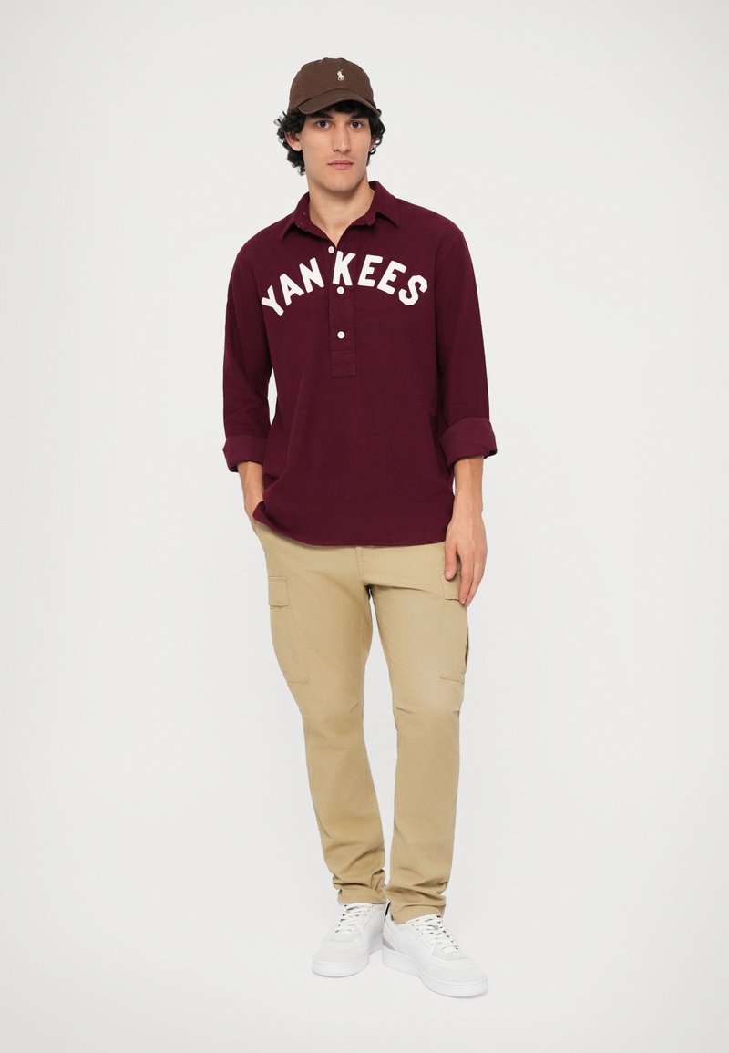 Maroon langermet polo med "YANKEES" skrift, knappelukking på kragen; kombinert med beige cargo-bukser og hvite sneakers. Brun caps.