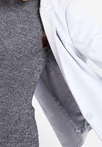 Grau strukturiertes Kleid mit seitlichem Ausschnitt, kombiniert mit einem weißen Blazer, der ein glattes Innenfutter und einen hellblauen Knopf hat.