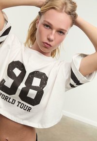 Witte cropped t-shirt met zwarte opdruk "98" en de tekst "WORLD TOUR". Heeft brede mouwen met zwarte strepen. Gemaakt van zachte katoen.