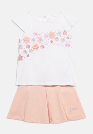 Top bianco a maniche corte con decorazioni floreali rosa e argento abbinato a una gonna svasata rosa chiaro con un piccolo logo "Guess".