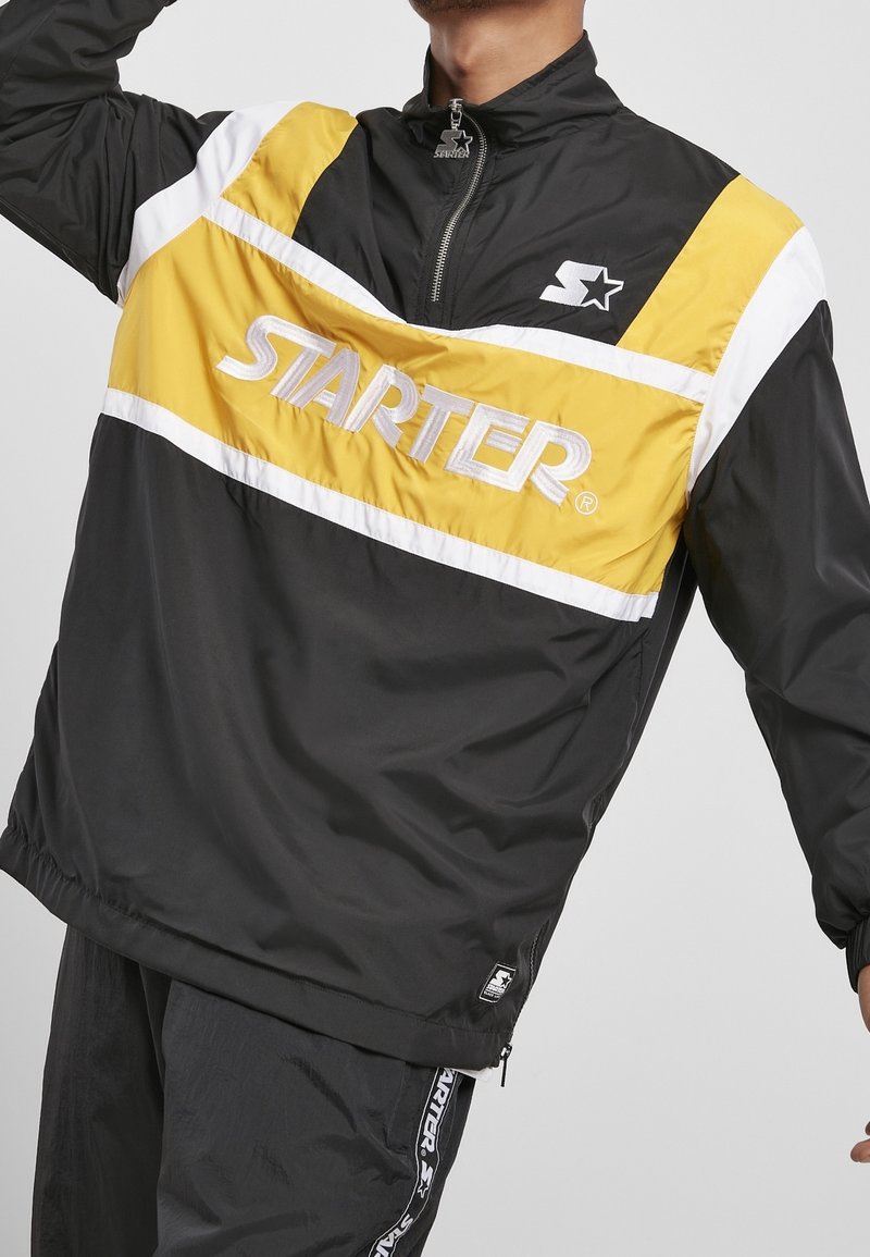 Homme portant une veste coupe-vent de la marque Starter noire, jaune et blanche avec un logo sur la poitrine et un pantalon de survêtement assorti.