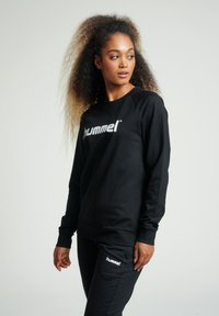 Felpa nera con maniche lunghe e polsini a coste, caratterizzata da un evidente logo bianco "hummel" sul petto, abbinata a pantaloni jogger coordinate.