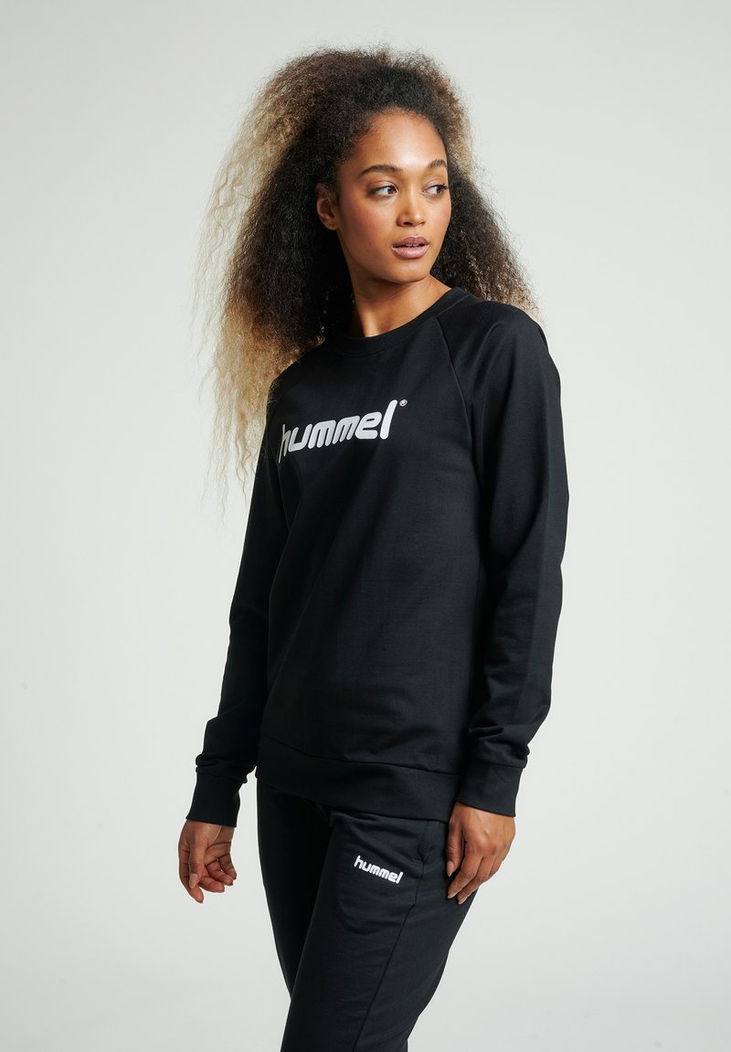 Felpa nera con maniche lunghe e polsini a coste, caratterizzata da un evidente logo bianco "hummel" sul petto, abbinata a pantaloni jogger coordinate.
