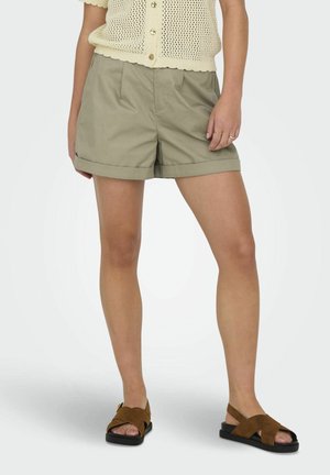 Mujer con top de punto beige, pantalones cortos caqui remangados, sandalias marrones con tiras cruzadas y anillos en la mano derecha, de pie contra un fondo liso.