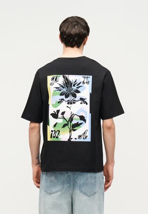 Személy fekete, túlméretezett pólót visel, melynek hátulján nagy, színes virágos grafika és stilizált szöveg található, világoskék hímzett farmerrel párosítva.