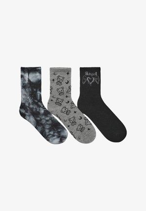 Trzy pary skarpetek: jedna w czarno-szary tie-dye, jedna szara z wzorem misia i księżyca, a jedna czarna z napisem "Anioł" i wzorem skrzydeł.