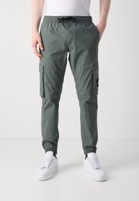 Lekkie zielone spodnie cargo z elastycznym pasem, bocznymi kieszeniami i ściągaczami. Stylizowane z białymi sportowymi butami dla kontrastu.