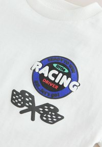 Chemise blanche avec un écusson circulaire de course bleu et noir portant la mention "little RACING DRIVER" et deux drapeaux à damier noirs croisés en dessous.