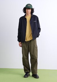 Black button-up jacket over a mustard shirt, paired with olive cargo pants and black shoes. Green cap adds detail to the outfit.