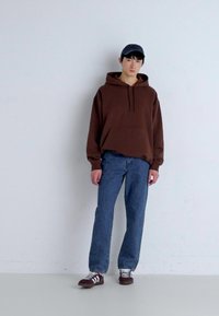 Brun bomullsgenser med frontlomme, kombinert med blå denimjeans. Modellen har på seg hvite og brune sneakers og en marineblå caps. Hvit bakgrunn.