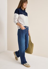 Felpa bianca e blu navy con il testo "UNIQUE", abbinata a jeans a gamba larga blu, sneakers beige e una borsa verde, su uno sfondo neutro.