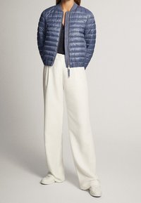 Femme debout portant une veste matelassée bleue, un haut noir, un pantalon blanc à jambes larges et des baskets blanches sur un fond uni.