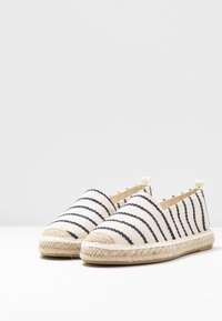 Canvas espadriller med marinblå ränder på en krämig bakgrund. Har en sula av jutesnöre och en flik vid hälen för enklare påtagning.