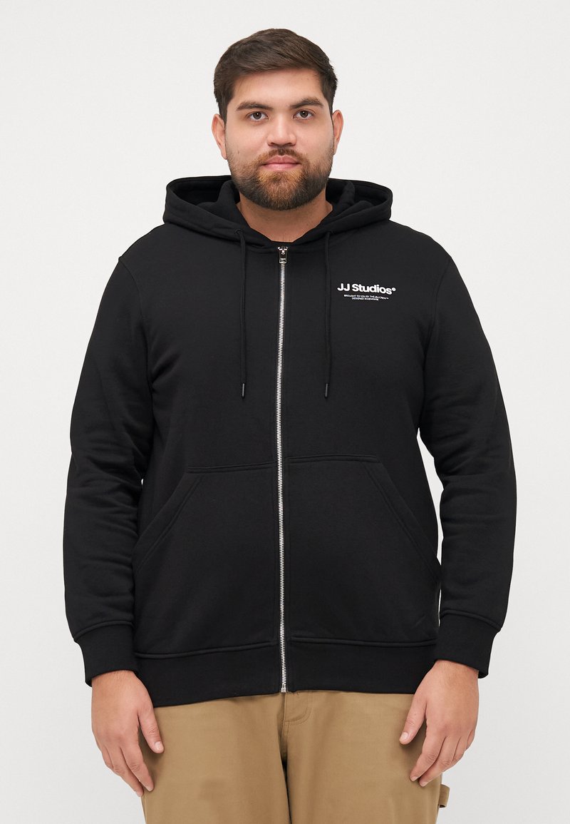 Zwarte zip-up hoodie met kangoeroezakken, capuchon met trekkoord en witte "JJ Studios" logo op de borst. Getextureerde stof met ribbels aan de manchetten.