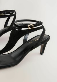 Zwarte suède hakken sandalen met kruislings straps en enkelgesp, met een slanke leren binnenzool en een taps toelopende hak.