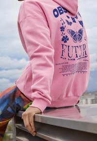 Sudadera rosa hecha de tela suave que presenta texto gráfico azul e ilustraciones de mariposas en la parte delantera, combinada con pantalones de estampado multicolor.