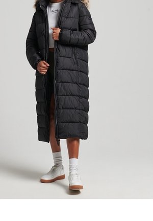 Personne portant un long manteau matelassé noir avec capuche en fourrure, chaussettes blanches, baskets blanches et un haut court blanc.
