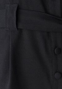 Juoda audinio medžiaga su lygiu paviršiumi, kurioje siūtas diržas ir aplink siūlę išdėstyti apvalūs juodi mygtukai.