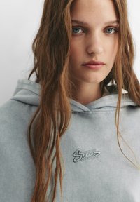 Haut de sweat gris clair avec une poche avant, présentant un logo brodé sur la poitrine. Texture douce et capuche avec cordon de serrage ajustable.