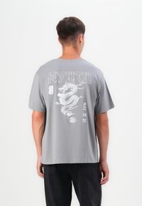 Grå kortærmet t-shirt med et dragegrafik p�å ryggen, der har hvid tekst og intrikate linjedetaljer, lavet af blødt bomuldsstof.