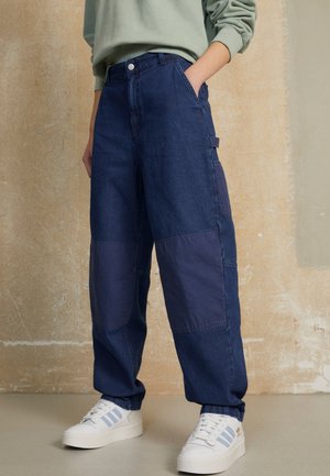 Person trägt weit geschnittene dunkelblaue Patchwork-Jeans, hellgrünes Sweatshirt und weiße Turnschuhe mit hellblauen Streifen, steht vor einer strukturierten Wand.