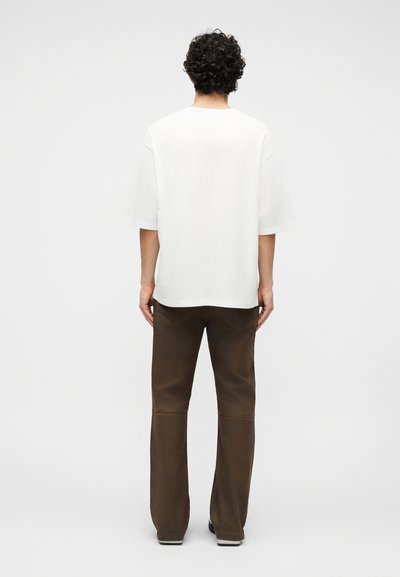 Άτομο με σγουρά μαλλιά, που στέκεται με την πλάτη γυρισμένη, φορά ένα άνετο λευκό πουκάμισο με κοντά μανίκια και καφέ παντελόνια με ίσια γραμμή, μπροστά σε απλό φόντο.