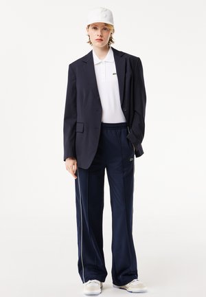 Blazer ajusté bleu marine sur un polo blanc, associé à un pantalon de survêtement bleu marine avec des rayures sur les côtés et une casquette blanche. Détail clé du logo sur le pantalon.