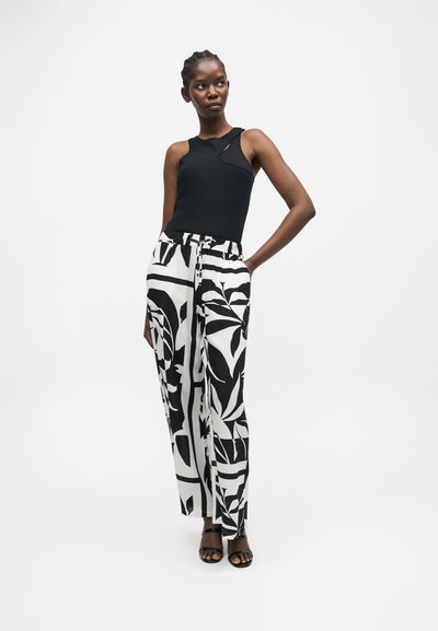 Γυναίκα που φοράει μαύρο αμάνικο τοπ με κοψίματα και ψηλόμεσο λευκό παντελόνι με έντονα μαύρα αφηρημένα φύλλα, ποζάρει με το χέρι στη μέση.