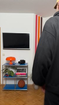 Fekete kapucnis pulóvert viselő személy egy fém polc mellett áll, amelyen narancssárga lámpa, kamera, könyvek, növények és kosárlabda találhatók, egy falra szerelt televízió és csíkos faldekoráció alatt.