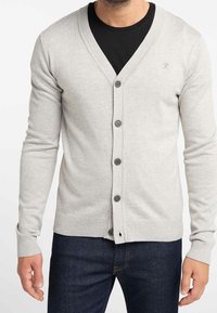 Cardigan a maniche lunghe, di un grigio chiaro, realizzato in tessuto a maglia. Design a V con sei bottoni e polsini e orlo a coste. Indossato sopra una maglietta nera.